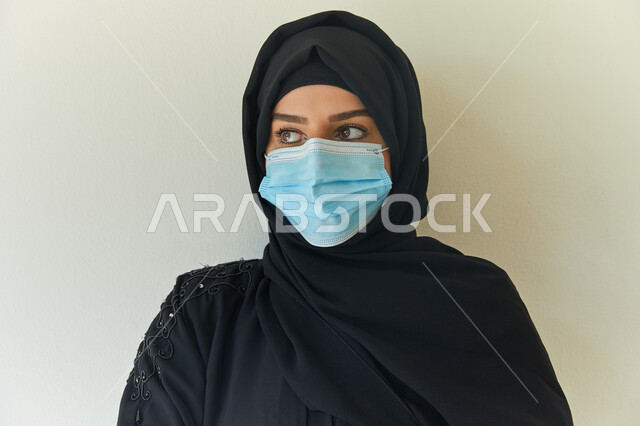 Follow the preventive and safety measures to protect from infection with Covid-19, gestures indicating looking at something, a close-up portrait of a veiled Arab Gulf Emirati woman wearing a black abaya and wearing a muzzle to avoid the spread of the Coro