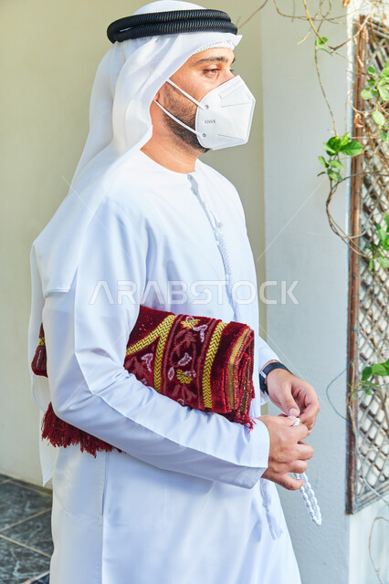 التسبيح والأذكار اليومية، رجل عربي خليجي اماراتي يرتدي الكندورة والغترة يحمل بيده السبحة وسجادة الصلاة، الاستعداد للذهاب وتأدية الصلوات في المسجد، العبادة و التقرب الى الله، الالتزام بالاجراءات الاحترازية وارتداء الكمامة للوقاية من الامراض وفايروس كورونا.