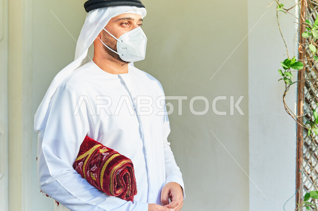 اسلاميات وعبادة، الاستعداد للذهاب وتأدية الصلوات في المسجد، التقرب الى الله بالتسبيح والأذكار اليومية، رجل عربي خليجي اماراتي يرتدي الكندورة والغترة يحمل بيده السبحة وسجادة الصلاة، الالتزام بالاجراءات الاحترازية وارتداء الكمامة للوقاية من فايروس كورونا.