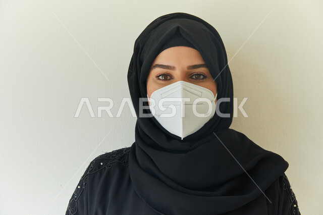 أخذ التدابير والإجراءات اللازمة لتجنب الإصابه بفايروس كورونا، بورتريه مقرب لامرأة عربية خليجية اماراتية محجبة ترتدي عباءه سوداء وتضع الكمامة لمنع العدوى بكوفيد- ١٩، إيماءات النظر إلى الكاميرا، خلفية بيضاء.