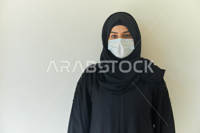 إتخاذ التدابير والإجراءات اللازمة لتجنب الإصابه بفايروس كورونا، بورتريه مقرب لامرأة عربية خليجية اماراتية محجبة ترتدي عباءه سوداء وتضع الكمامة لمنع العدوى بكوفيد- ١٩، إيماءات النظر إلى الكاميرا، خلفية بيضاء.
