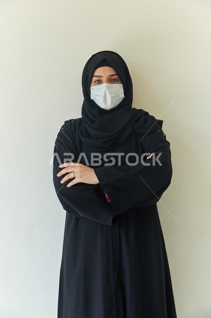 إحترام قوانين وقواعد وزاره الصحة لتجنب الاصابه بكوفيد-١٩، بورتريه لامرأة عربية خليجية اماراتية محجبة ترتدي عباءه سوداء وتضع الكمامة لتجنب تفشي فايروس كورونا، إيماءات النظر إلى الكاميرا ويدين متقاطعه، خلفية بيضاء.