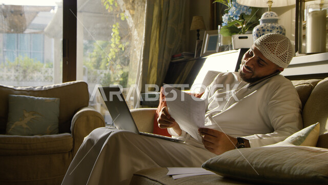 إنجاز مهام العمل عبر جهاز الحاسوب، رجل عربي خليجي إماراتي يرتدي الكندورة والطاقية الاماراتية البيضاء يقوم بإجراء مكالمة عبر الجوال، العمل عن بعد في زمن الكورونا واتباع طرق الوقاية، مفهوم ادارة الأعمال وعقد الاجتماعات عبر الانترنت