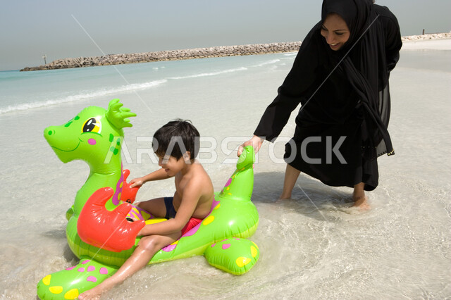 صبي يجلس على عوامة السباحة ويستمتع بالفعاليات الصيفية رفقة والدته, أجواء أسرية ممتعة, أم عربية إمراتية خليجية تقوم باللعب على شاطئ البحر مع ابنها في رحلة صيفية, أنشطة ترفيهية عائلية في العطلة, منتجعات شاطئية في الإمارات