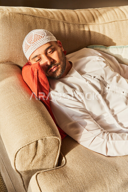 شرود الذهن والنظر إلى شيء ما، رجل عربي خليجي اماراتي يرتدي الكندورة والطاقية الاماراتية البيضاء يقوم بالاستلقاء على الاريكة في منزله في وقت النهار، اخذ قسط من الراحة، التفكير والحيرة في التخطيط للمستقبل، حل المشكلات واتخاذ القرارات.
