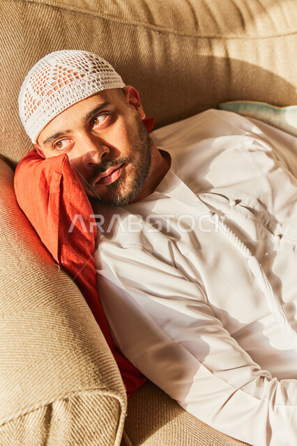 التفكير والحيرة في التخطيط للمستقبل، حل المشكلات واتخاذ القرارات، رجل عربي خليجي اماراتي يرتدي الكندورة والطاقية الاماراتية البيضاء يقوم بالاستلقاء على الاريكة في منزله في وقت النهار، اخذ قسط من الراحة، شرود الذهن والنظر إلى شيء ما.