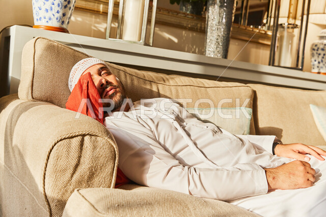 النوم بعمق من الارهاق والتعب، رجل عربي خليجي اماراتي يرتدي الكندورة والطاقية الاماراتية البيضاء يقوم بالاستلقاء على الاريكة في منزله، اخذ قيلولة و قسط من الراحة، الكسل والخمول والحاجة الى النوم. 