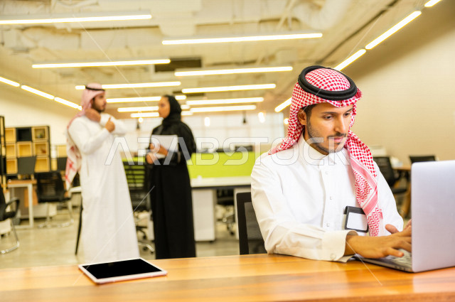 رجل اعمال سعودي خليجي على مكتب و يعمل على الكمبيوتر المحمول ، بالخلفية مجموعة رياديون من رجال و سيدات الأعمال السعوديون , شركة سعودية ، شركاء عمل ، بيئة عمل ، وظيفة مكتبية ، عمل خليجي ،  فريق العمل 