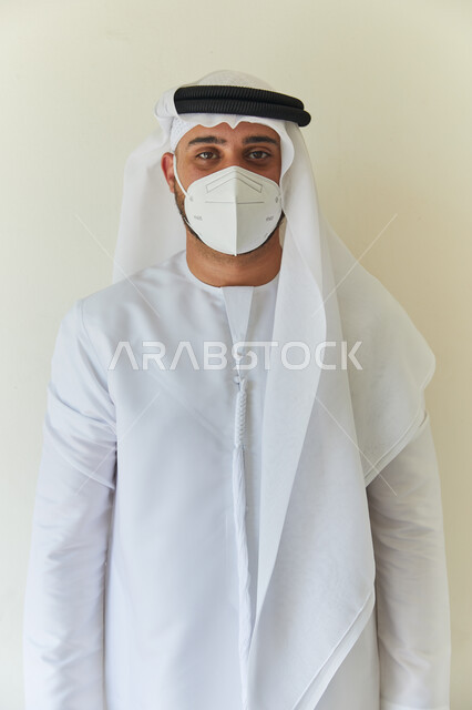 Avoiding surface contact and maintaining distances, following preventive measures against viruses, a close-up portrait of an Emirati Arab man wearing a kandora and a ghutra wearing a protective medical mask to protect from the Corona virus, a white backgr