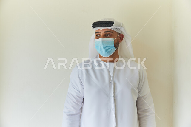 إتباع الإجراءات الوقائيه ضد الفايروسات، بورتريه مقرب لرجل عربي خليجي اماراتي يرتدي الكندوره والغتره يضع القناع الطبي الوقائي للوقايه من فايروس كورونا، الحفاظ على المسافات وتجنب الملامسه السطحيه، خلفيه بيضاء.