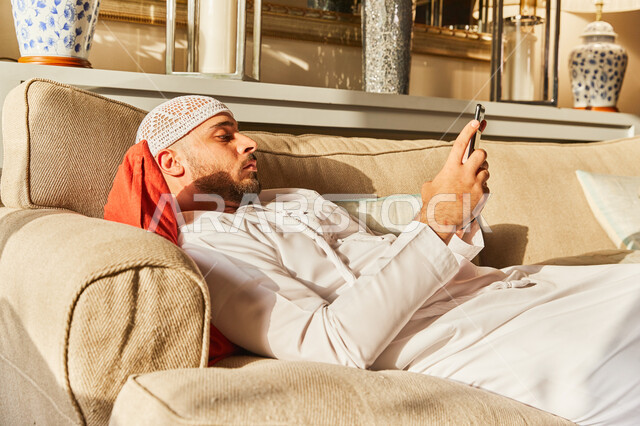 The concept of e-marketing, an Arab Gulf Emirati man wearing a kandora and a white Emirati cap lying on the sofa in his house holding a mobile phone in his hands, browsing websites and social media, spending fun times in online games via mobile phone.