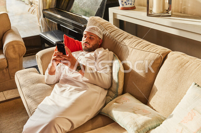 تصفح مواقع ووسائل التواصل الاجتماعي، مفهوم التسويق الالكتروني، رجل عربي خليجي اماراتي يرتدي الكندورة والطاقية الاماراتية البيضاء يستلقي على الأريكة في منزله يحمل الجوال بيديه، قضاء اوقات ممتعة في العاب الاونلاين عبر الهاتف المحمول. 