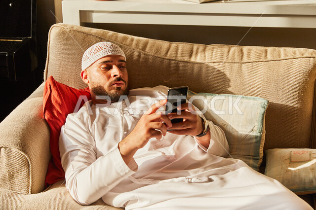 تصفح مواقع ووسائل التواصل الاجتماعي، مفهوم التسويق الالكتروني، رجل عربي خليجي اماراتي يرتدي الكندورة والطاقية الاماراتية البيضاء يستلقي على الأريكة في منزله يحمل الجوال بيديه، قضاء اوقات ممتعة في العاب الاونلاين عبر الهاتف المحمول. 