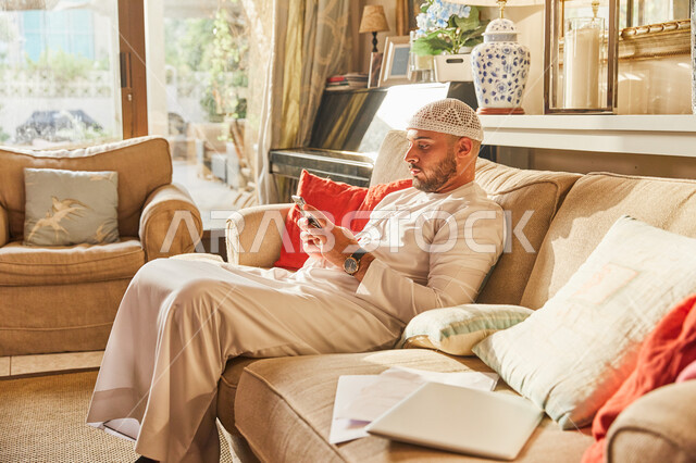 قضاء اوقات ممتعة في العاب الاونلاين عبر الهاتف المحمول، تصفح مواقع ووسائل التواصل الاجتماعي، رجل عربي خليجي اماراتي يرتدي الكندورة والطاقية الاماراتية البيضاء يجلس على الأريكة في منزله يحمل الجوال بيديه، مفهوم التسويق الالكتروني. 