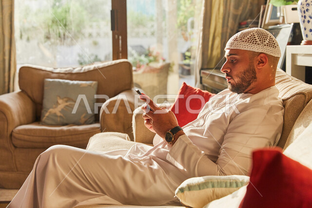 قضاء اوقات ممتعة في العاب الاونلاين عبر الهاتف المحمول، تصفح مواقع ووسائل التواصل الاجتماعي، رجل عربي خليجي اماراتي يرتدي الكندورة والطاقية الاماراتية البيضاء يجلس على الأريكة في منزله يحمل الجوال بيديه، مفهوم التسويق الالكتروني. 