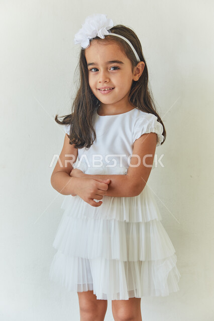 الإهتمام بالمظهر الخارجي للأطفال الصغار، بورتريه لطفلة عربيه خليجيه إماراتية تنظر إلى الكاميرا بإبتسامه لطيفه مكتفه اليدين وترتدي فستان أبيض اللون، ارتداء الملابس الجديده في الأعياد والمناسبات، خلفيه بيضاء