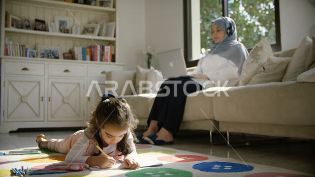 التسويق الإلكتروني في المنزل, تنمية القدرات والمهارات الفكرية، امرأه عربيه اماراتية خليجيه محجبه تجلس في غرفة المعيشة  تضع سماعات الهيدفون وتقوم بالعمل على جهاز الحاسوب المحمول بإيماءات السعادة، فتاة عربية اماراتية تقوم بنشاط التلوين ، العمل عن بعد