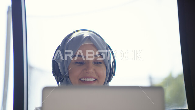 التسلية والترفيه وقضاء أوقات ممتعه في أيام العطلة، إجراء مكالمة فيديو عبر الحاسوب المحمول، صورة مقربة لإمرأة عربيه إماراتية خليجية محجبه مبتسمة تجلس في غرفة المعيشة ترتدي سماعات الرأس الصوتية، التواصل مع الأصدقاء بإيماءات السعادة، استخدام تقنية الهيدفون
