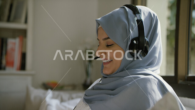 الاستمتاع في الإصغاء إلى المقاطع الغنائية، صورة مقربة لإمرأة عربيه إماراتية خليجية محجبه مبتسمة  تجلس في غرفة المعيشة ترتدي سماعات الرأس الصوتية، الاستمتاع بسماع الموسيقى بإيماءات السعادة، استخدام تقنية الهيدفون، التسلية والترفيه وقضاء أوقات ممتعه