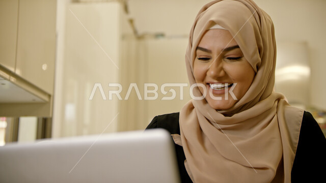 التواصل الاجتماعي مع الاصدقاء ،صورة مقربة لسيدة عربية إماراتية خليجية محجبه مبتسمة تقوم بإجراء مكالمة فيديو عبر الحاسوب المحمول بإيماءات السعادة، التسويق الإلكتروني في المنزل، العمل عن بعد، تصفح الانترنت واستخدام تقنيات حديثة