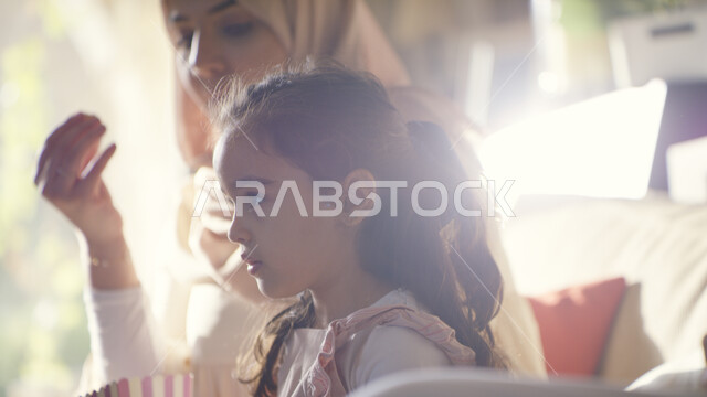 قضاء اوقات ممتعه مع الأبناء في العطلة الصيفية، الاستمتاع بأوقات الفراغ وتناول الفوشار, صورة مقربة لفتاة عربية اماراتية خليجية تجلس مع عائلتها في غرفة المعيشة وتقوم بمشاهدة البرامج المفضلة على التلفاز، جلسات عائلية مميزة 