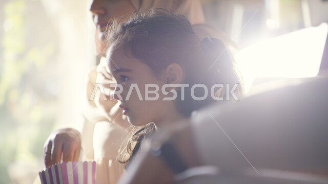  الاستمتاع بأوقات الفراغ وتناول الفوشار, صورة مقربة لفتاة عربية اماراتية خليجية تجلس مع عائلتها في غرفة المعيشة وتقوم بمشاهدة البرامج المفضلة على التلفاز، قضاء اوقات ممتعه مع الأبناء في العطلة الصيفية، جلسات عائلية مميزة 