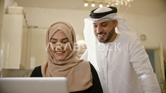 التسويق الإلكتروني في المنزل, أخبار جديده على وسائل الإعلام ومواقع التواصل الاجتماعي ،صورة مقربة لسيدة عربية إماراتية خليجية محجبه يقف وراءها زوجها تقوم بتصفح الانترنت واستخدام تقنيات حديثة، إجراء مكالمة فيديو عبر الحاسوب المحمول بإيماءات السعادة