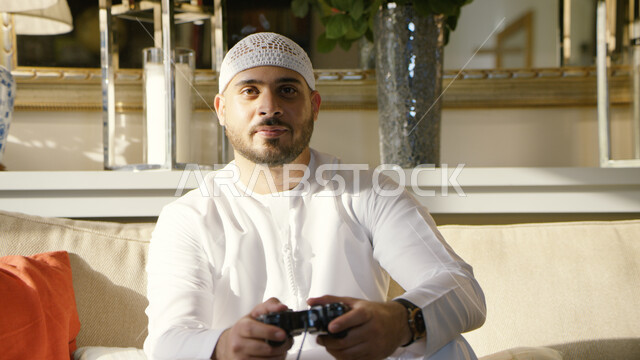 المتعة و التسلية بألعاب البلايستيشن الترفيهيه، شاب عربي اماراتي خليجي يرتدي الطاقية البيضاء والكندورة يحمل بيده عصا التحكم ويلعب بألعاب الفيديو،  قضاء أوقات ممتعه في المنزل، التقنيات الحديثة والتكنولوجيا، الاستمتاع بأوقات الفراغ في العطلة الصيفية