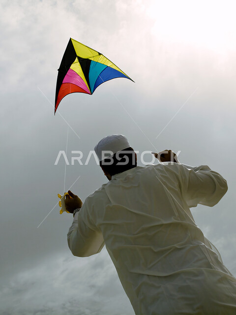 السياحة في دول الخليج العربي والاستمتاع بالاجازة الصيفية، الإستجمام والتنزه في الهواء الطلق, صورة من الخلف لرجل عربي خليجي إماراتي يرتدي الثوب الخليجي يطير طائرة ورقية على الشاطئ, قضاء يوم العطلة على البحر