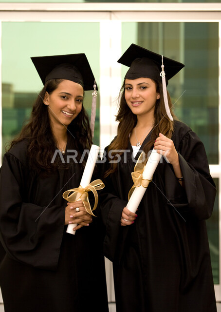 ايماءات وجه تدل على السعادة والفرح والفخر، التعليم في جامعات الإمارات العربية، الاحتفال بالتخرج من الجامعة، صورة مقربة لخريجتين جامعيتين عربيتين خليجيتين إماراتيتين ترتديان ملابس التخرج وتمسكان شهادة النجاح والتفوق.
