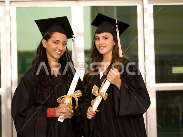 التعليم في جامعات الامارات العربية، الاحتفال بالتخرج من الجامعة، صورة مقربة لخريجتين جامعيتين عربيتين خليجيتين اماراتيتين ترتديان ملابس التخرج وتمسكان شهادة النجاح والتفوق، ايماءات وجه تدل على السعادة والفرح والفخر.