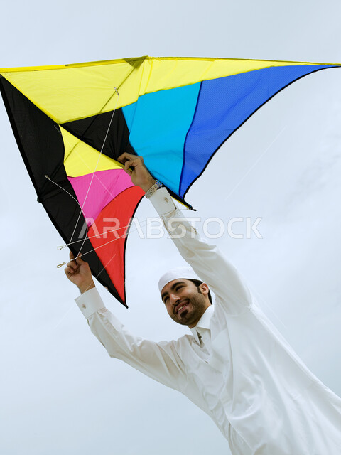 Gesture of two hands indicating enthusiasm and joy, tourism in the Arab Gulf countries and enjoying the summer vacation, recreation and hiking in the open air, an Arab Emirati man wearing a Gulf dress flying a kite on the beach