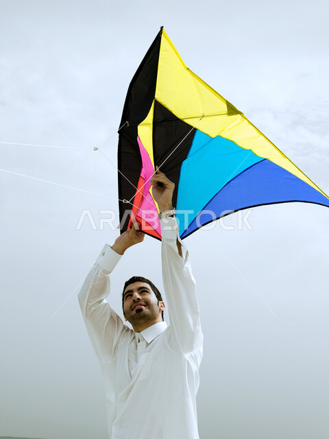 Recreation and hiking in the open air, hand gestures indicating enthusiasm and joy, tourism in the Arab Gulf countries and enjoying the summer vacation, an Emirati Arab man wearing a Gulf dress flying a kite on the beach