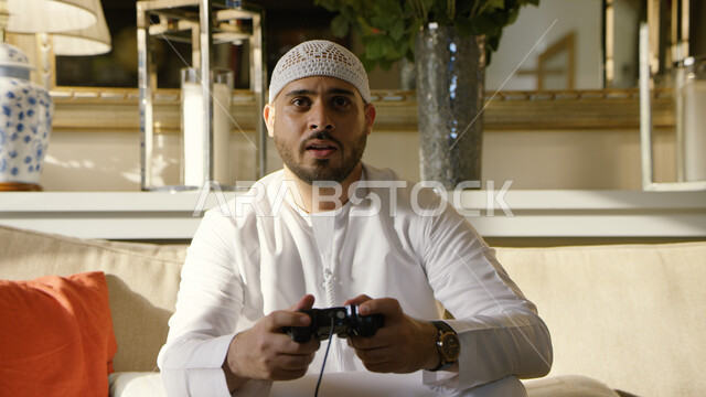   قضاء أوقات ممتعه في المنزل، المتعة و التسلية بألعاب البلايستيشن الترفيهيه، شاب عربي اماراتي خليجي يرتدي الطاقية البيضاء والكندورة يحمل بيده عصا التحكم ويلعب بألعاب الفيديو، التقنيات الحديثة والتكنولوجيا، الاستمتاع بأوقات الفراغ في العطلة الصيفية