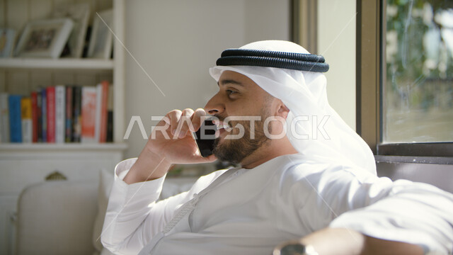 Gestures indicating happiness, making a voice phone call on a mobile phone, a close-up of an Arab Emirati Gulf man wearing an Emirati kandora and ghutra sitting in the living room holding a mobile phone in his hand, communicating with friends and relative
