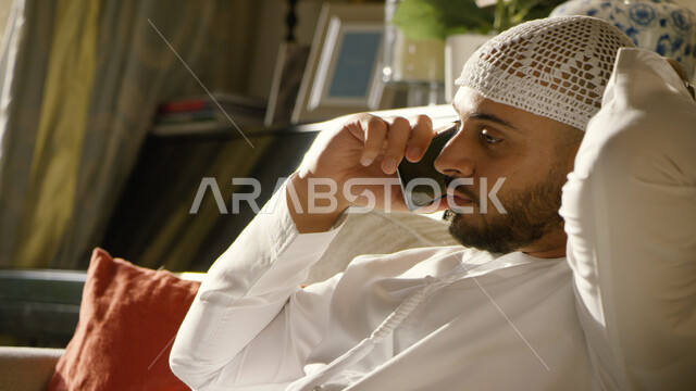 التواصل مع الأقارب والاصدقاء، إجراء مكالمة هاتفية صوتية عبر الجوال، صورة مقربة لرجل عربي اماراتي خليجي يرتدي الكندورة والطاقية البيضاء  يجلس في غرفة المعيشة ويحمل بيده الهاتف المحمول، ايماءات تدل على السعادة