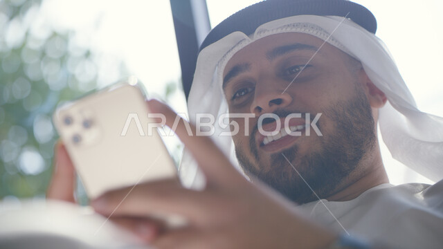 دردشات ومحادثات عبر مواقع التواصل الاجتماعي, صورة مقربة لرجل عربي خليجي إماراتي مبتسم يرتدي الكندورة والغترة يستخدم الهاتف المحمول, التواصل مع الأصدقاء عبر الانترنت بإيماءات الفرح والسرور
