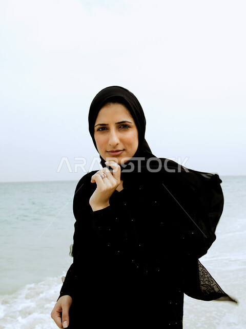 ايماءات وجه ويدين مختلفة, إمرأة عربية خليجية اماراتية محجبة ترتدي العباءة السوداء و تقف على شاطئ البحر، السياحة في دول الخليج العربي والاستمتاع بالاجازة الصيفية، الإستجمام والتنزه في الهواء الطلق