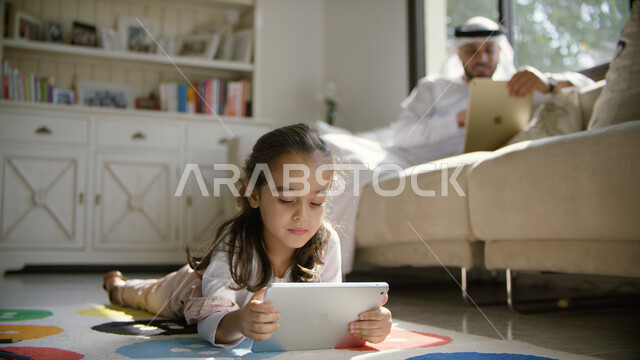 الترابط الأسري والمشاركة مع الأولاد النشاطات الترفيهيه، استخدام أجهزة التابليت في المنزل والاستفادة من أوقات الفراغ, أب عربي خليجي إماراتي يرتدي الكندورة والغترة يجلس في غرفة المعيشة برفقه ابنته, الألعاب التعليمية والتسليه عبر الانترنت مع الأبناء