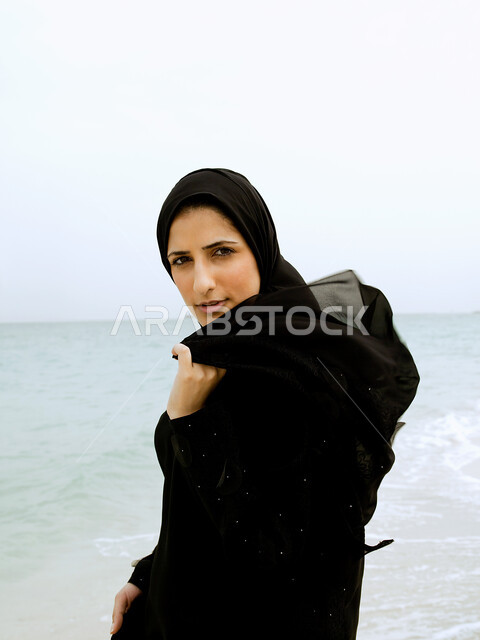 السياحة والاستمتاع بالاجازة الصيفية في دول الخليج العربي، ايماءات وجه ويدين مختلفة, إمرأة عربية خليجية اماراتية محجبة ترتدي العباءة السوداء و تقف على شاطئ البحر، الإستجمام والتنزه في الهواء الطلق