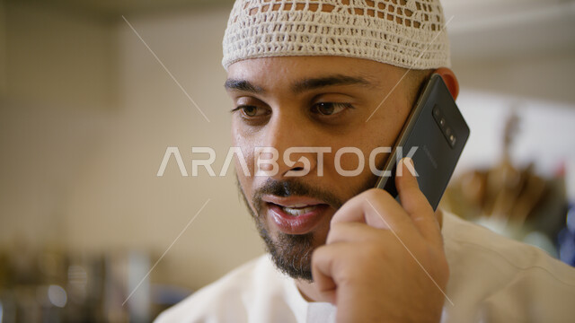دردشة ومحادثات مع الأقارب والاصدقاء، إجراء مكالمة هاتفية صوتية عبر الجوال، صورة مقربة لرجل عربي اماراتي خليجي يرتدي الكندورة والطاقية البيضاء يجلس في غرفة المعيشة ويحمل بيده الهاتف المحمول، ايماءات تدل على السعادة