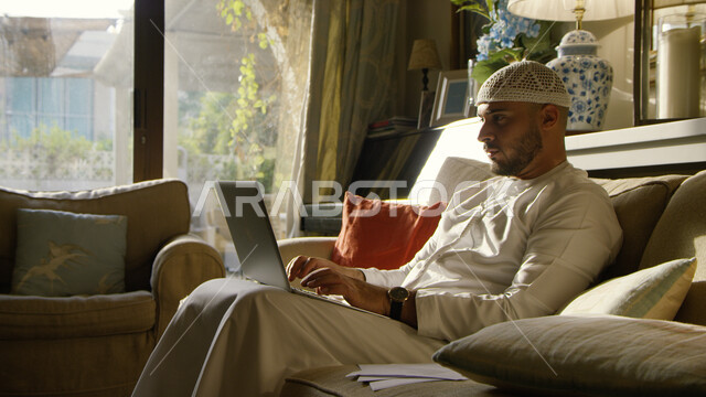 الجلوس في غرفة المعيشة وإنجاز مهام العمل عبر الانترنت في زمن الكورونا, رجل عربي خليجي إماراتي يرتدي الطاقية البيضاء والكندورة الاماراتية يستخدم جهاز الحاسوب في وقت الظهيرة, استخدام التطبيقات والتقنيات الحديثة لإدارة الأعمال عن بعد، النظر إلى شيء ما بإيماء