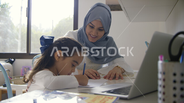 الاستعانة بمواقع الانترنت للحصول على أجوبة موثوقه، متابعه حصص الاونلاين عبر الحاسوب المحمول ،سيدة عربية اماراتية خليجية محجبة تقووم في مساعدة ابنتها في حل الواجب، الاهتمام والاعتناء بالأبناء، التعليم المنزلي اثناء الحجر الصحي و تفشي فيروس كورونا
