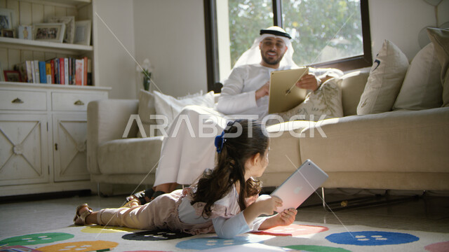 استخدام أجهزة التابليت في المنزل والاستفادة من أوقات الفراغ, أب عربي خليجي إماراتي مبتسم يرتدي الكندورة والغترة يجلس في غرفة المعيشة برفقه ابنته, الألعاب التعليمية والتسليه عبر الانترنت مع الأبناء, الاستمتاع بالأجواء العائلية الممتعة مع الأهل