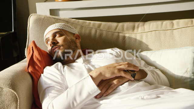 النظر إلى شيء ما وشرود الذهن, رجل عربي خليجي اماراتي يرتدي الكندورة والطاقية الاماراتية البيضاء يقوم بالاستلقاء على الأريكة في منزله وقت الظهيرة, أخذ قسط من الراحة, التفكير والحيرة في التخطيط للمستقبل, حل المشكلات واتخاذ القرارات