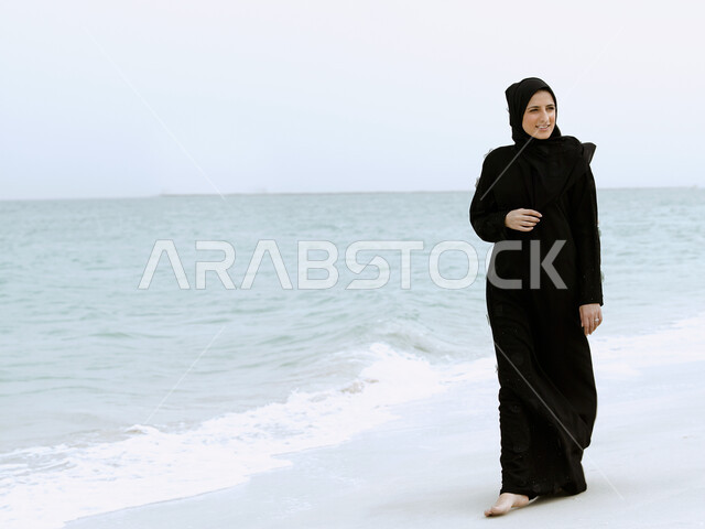 صورة الجسم بالطول الكامل, التنزه والترفيه في أجواء إماراتية سعيده, السياحة والاستمتاع بالاجازة الصيفية في دول الخليج العربي, إمرأة عربية خليجية اماراتية محجبة مبتسمة ترتدي العباءة السوداء تقف على ساحل البحر, قضاء رحلة سياحية على الشاطئ