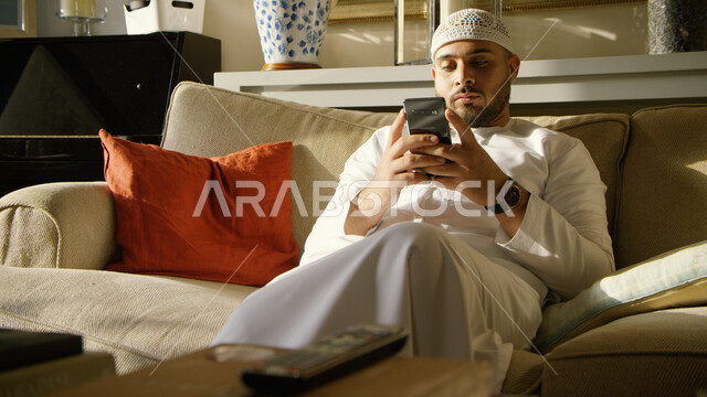 إرسال الرسائل النصية والتواصل مع الأصدقاء والأقارب في وقت النهار, متابعه الأخبار الجديدة على وسائل الاعلام بإيماءات الهدوء والملل، صورة مقربه لشاب عربي إماراتي خليجي يرتدي الكندورة والطاقية البيضاء مستلقي على الأريكة ويحمل بيده الهاتف المحمول