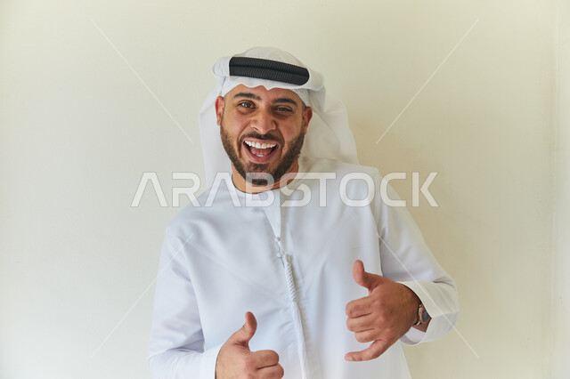 ايماءات وجه ويدين مختلفه تدل على الحماس والسعاده، الإعتناء بالمظهر الخارجي، بورتريه مقرب لشاب عربي خليجي إماراتي يرتدي الكندوره والغتره الإماراتيه يقوم بالنظر إلى الكاميرا، خلفيه بيضاء. 