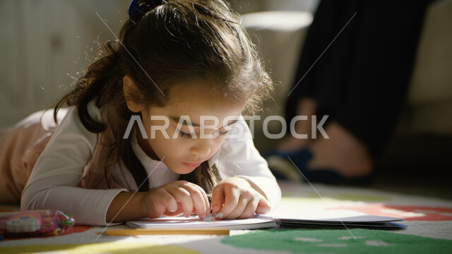 العودة إلى المدارس وبداية سنة دراسية جديدة, صورة مقربة لطفلة عربية خليجية إمارااتية تجلس على الأرض تقوم بحل الواجبات والدروس, متابعة الأولاد في المنزل وتنمية المهارات والابتكارات لدى الأبناء