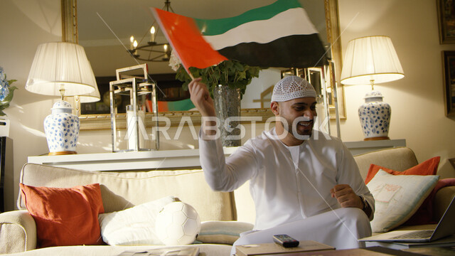 Passion in following the UAE national team with gestures of joy and achieving victory, watching football matches on the computer, an Arab Emirati man sitting in the living room wearing a white cap and a kandora holding the flag of the United Arab Emirates